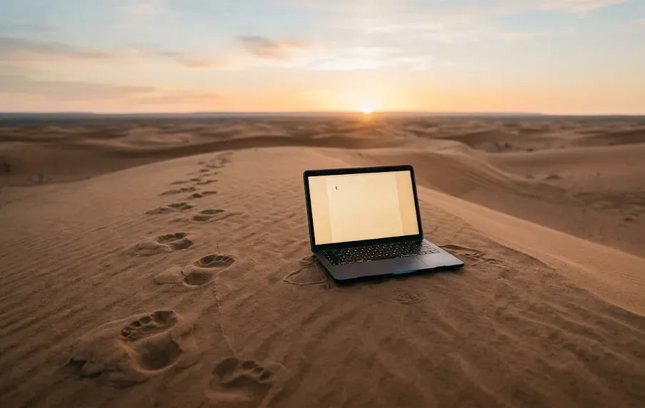 Una notebook con la pantalla en blanco, sola en medio de un desierto al amanecer, con huellas de pisadas que se detienen frente a ella